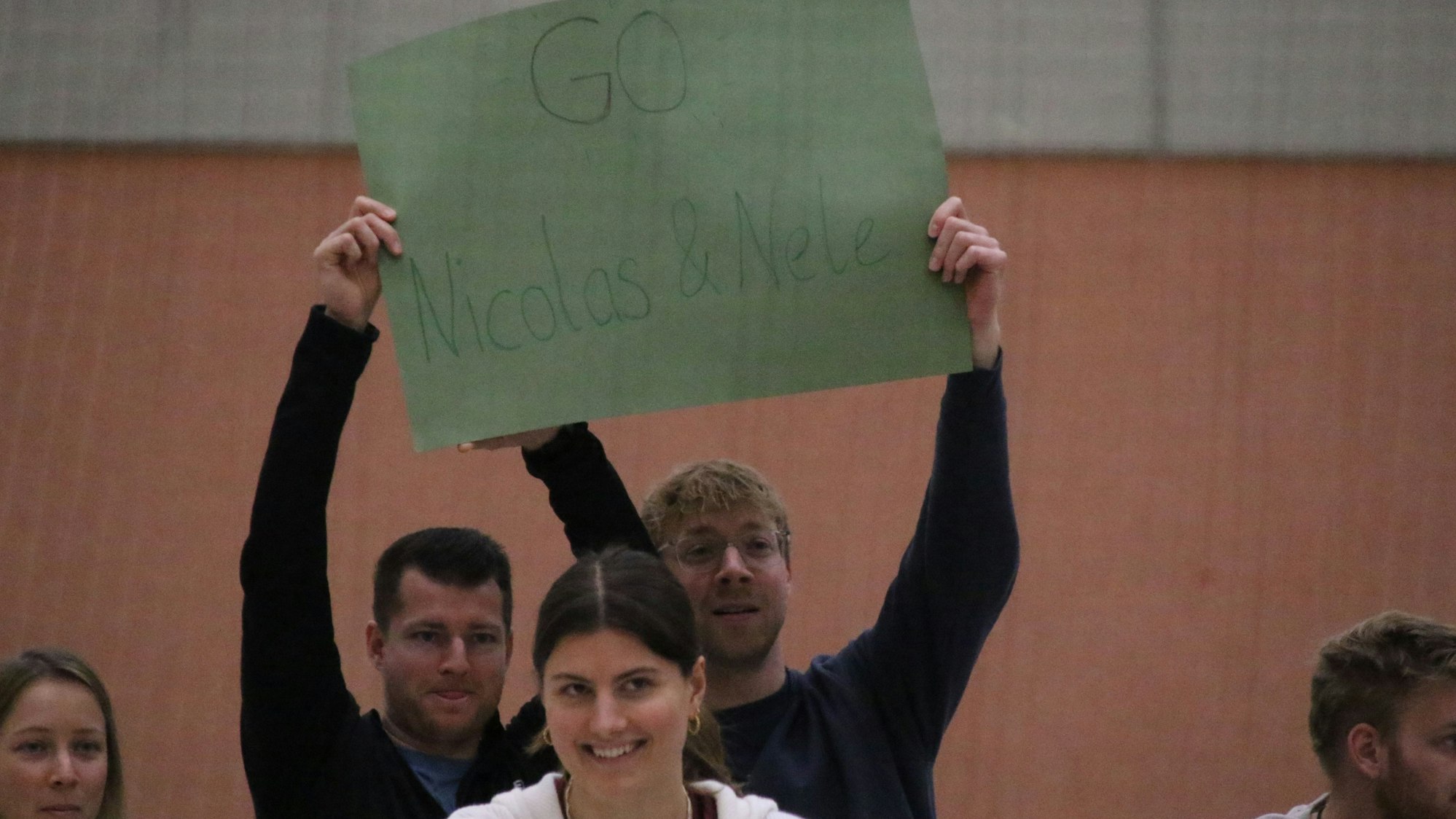 Mit einem grünen Pappschild feuern zwei Männer einen Spieler und eine Spielerin an.