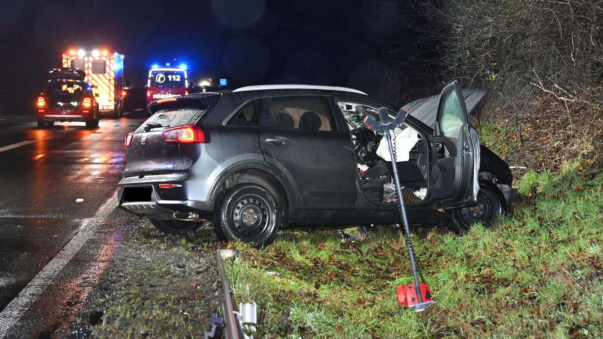 Ein verbeultes Auto steht neben einer regennassen Straße, im Hintergrund Einsatzfahrzeuge von Polizei und Feuerwehr.