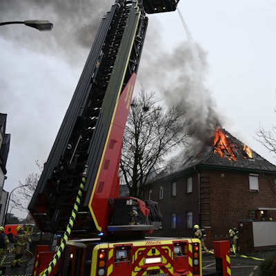 Ein Dachstuhl brennt im Hintergrund, ein Fahrzeug der Feuerwehr und Feuerwehrleute stehen im Vordergrund.