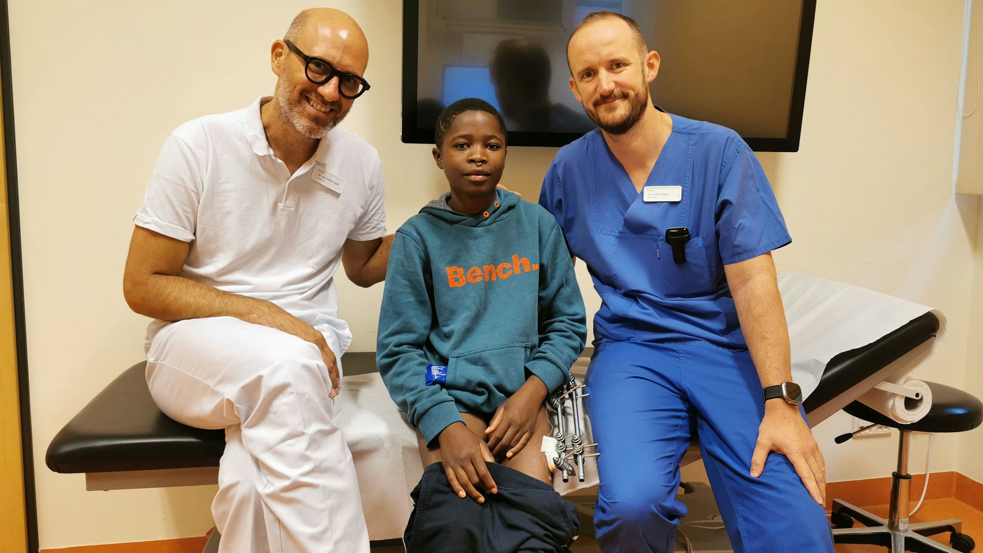 Zwei Ärzte sitzen mit einem dunkelhäutigen Jungen auf einer Behandlungsliege in einem Krankenhaus