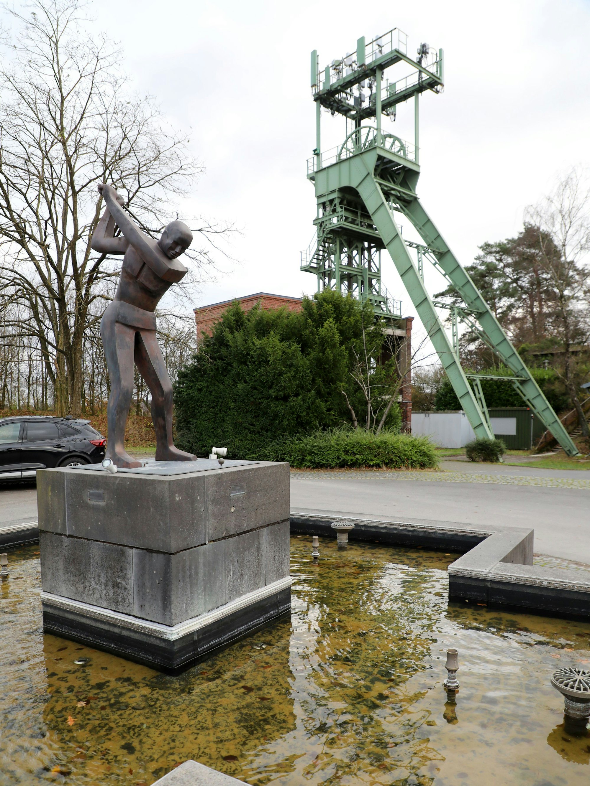 Auf einem Brunnen steht eine Golfspieler-Figur, im Hintergrund ist der historische Förderturm des Hauptschachts der Erzgrube Lüderich zu sehen.