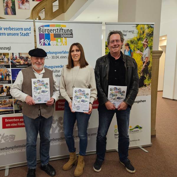Zwei Männer und eine Frau posieren mit Heften, die sie vor sich halten, vor einer Pappaufstellwand der Bürgerstiftung Schleiden.