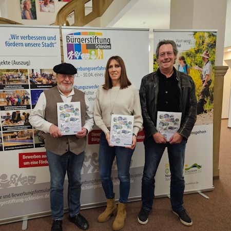 Zwei Männer und eine Frau posieren mit Heften, die sie vor sich halten, vor einer Pappaufstellwand der Bürgerstiftung Schleiden.