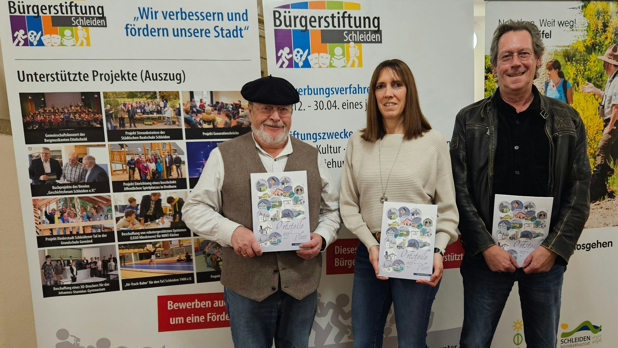 Zwei Männer und eine Frau posieren mit Heften, die sie vor sich halten, vor einer Pappaufstellwand der Bürgerstiftung Schleiden.