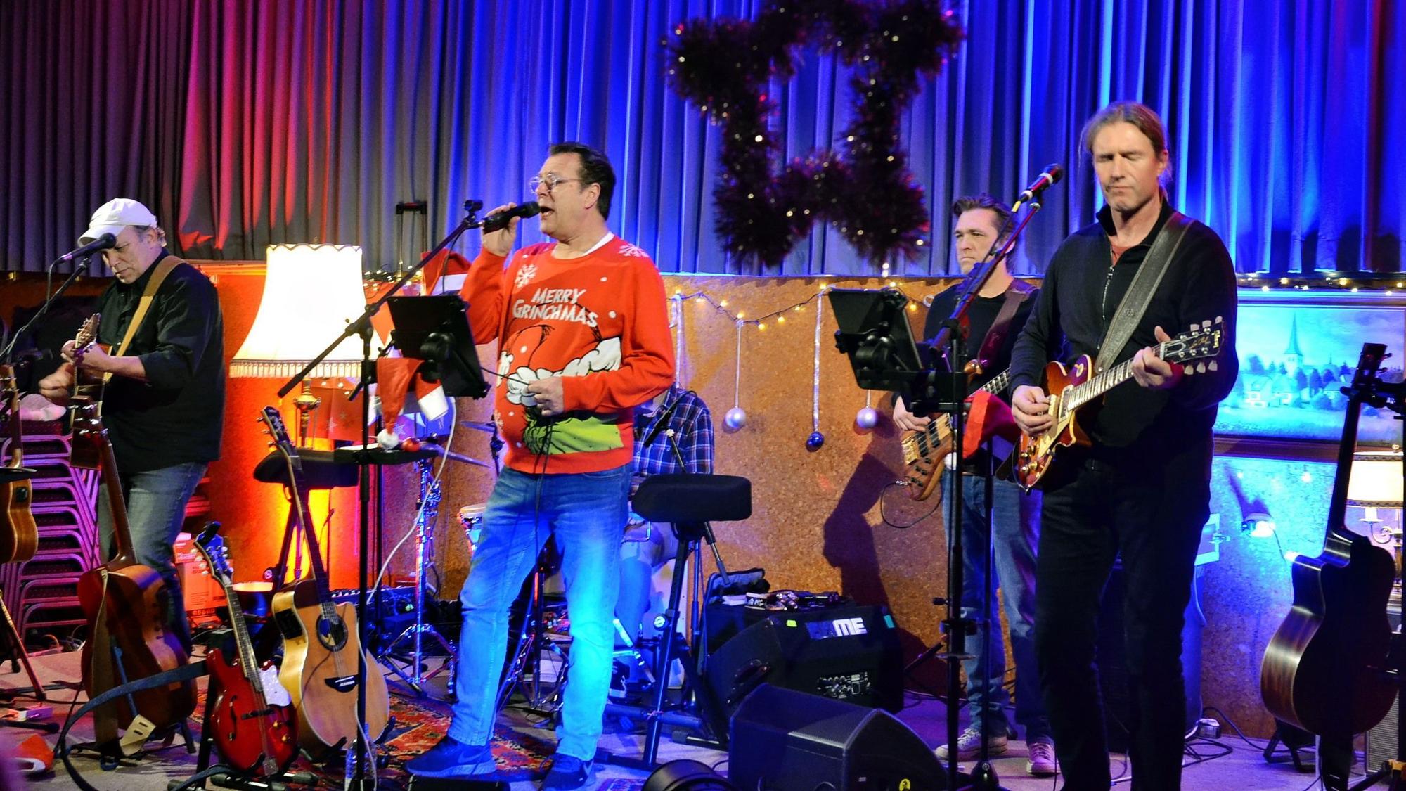 Ein Sänger im roten Weihnachtspullover steht an einem Mikrofon, um ihn herum die anderen Musiker der Band „Rock on Wood“.