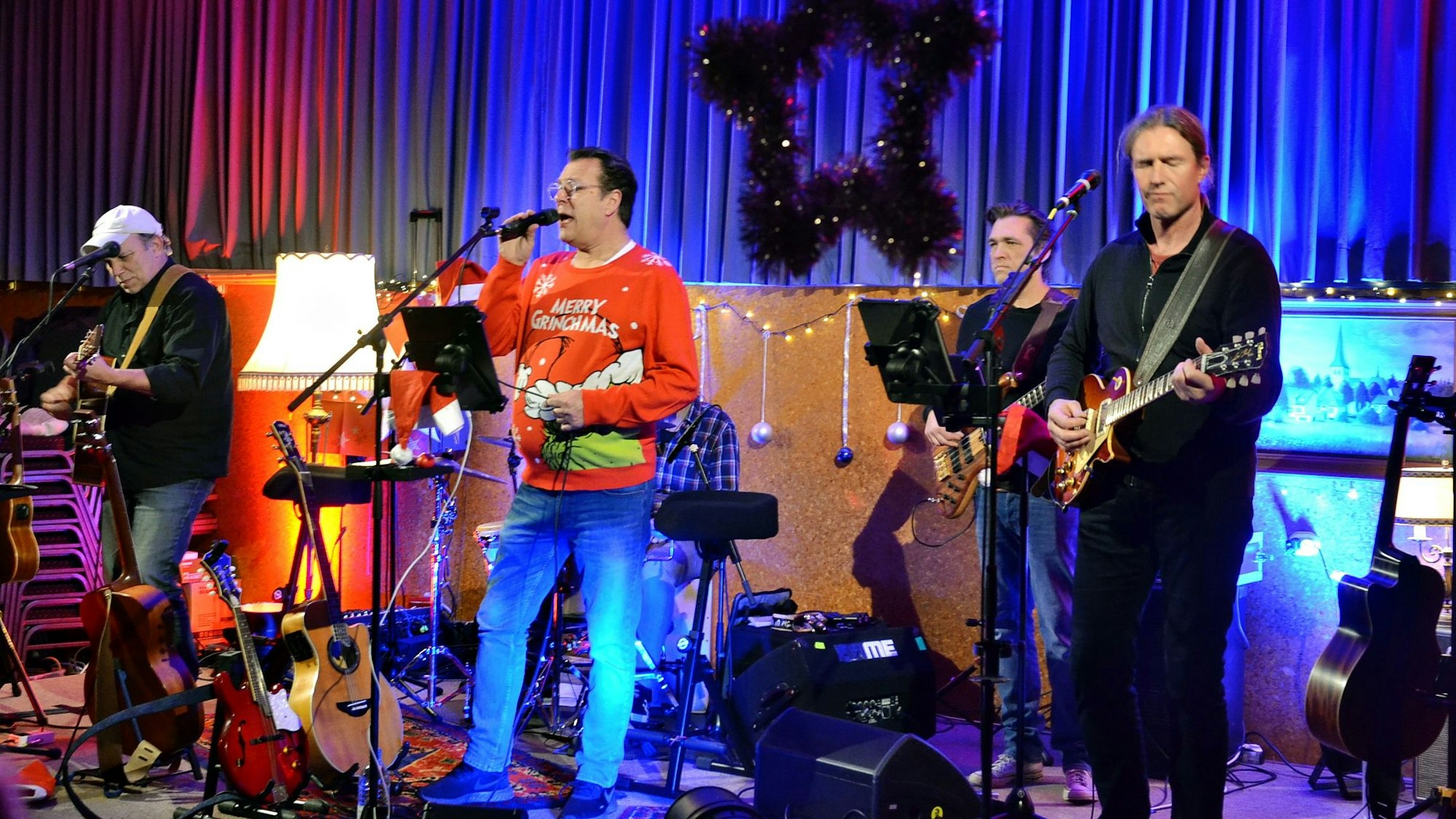 Ein Sänger im roten Weihnachtspullover steht an einem Mikrofon, um ihn herum die anderen Musiker der Band „Rock on Wood“.