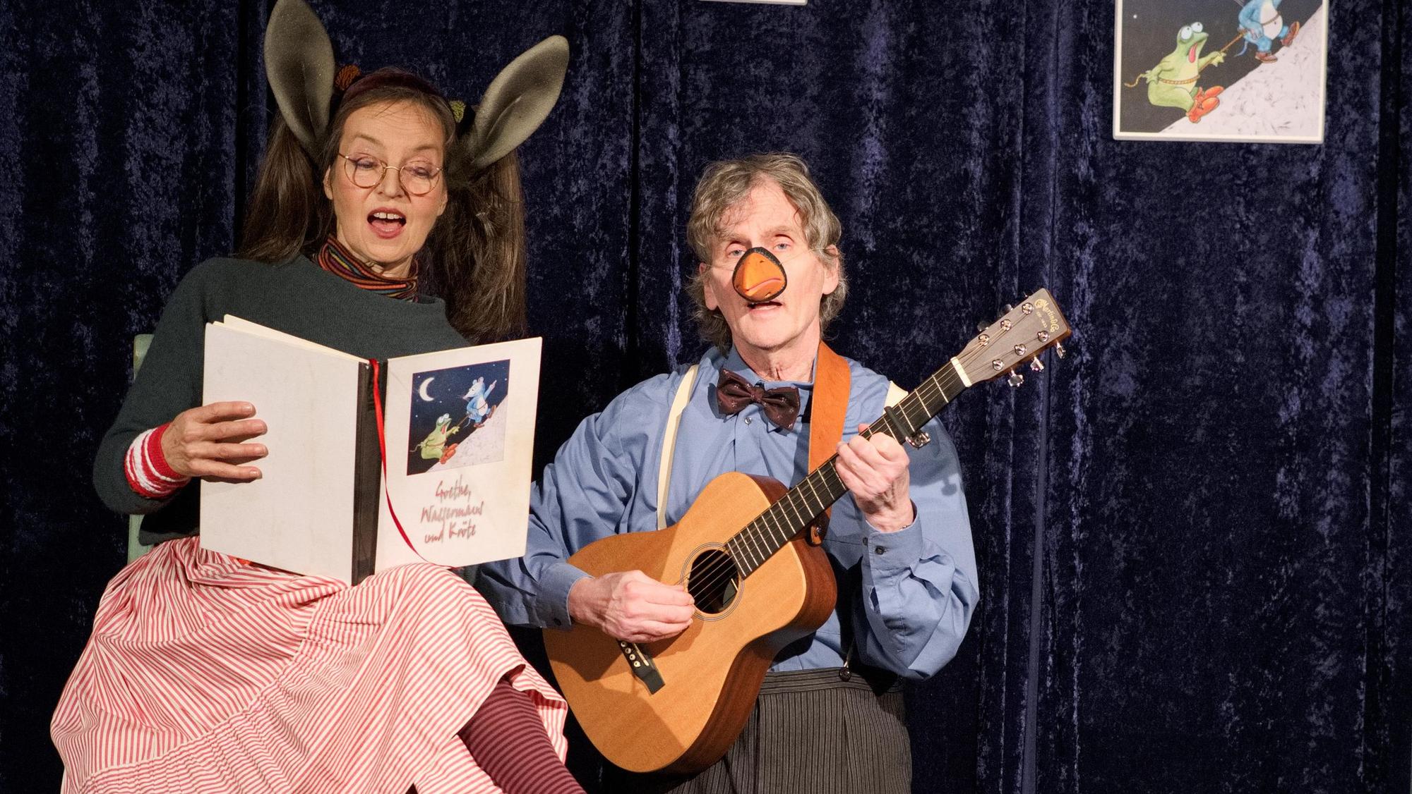 Christiane Remmert, ausstaffiert mit Hasenohren, liest aus einem Buch, während Jojo Ludwig sie mit der Gitarre begleitet.