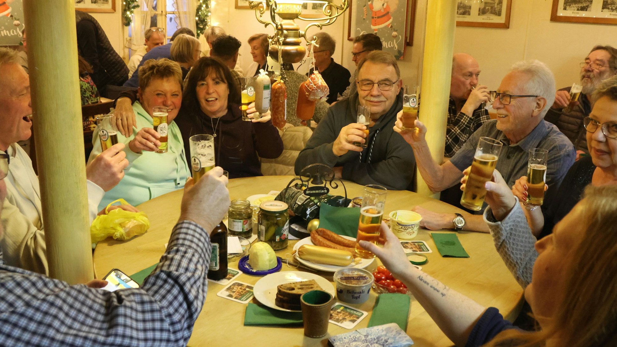 Zu sehen sind Menschen, die in einer Kneipe an einem Tisch ein Würfelspiel spielen. An der Lampe hängen Würste.