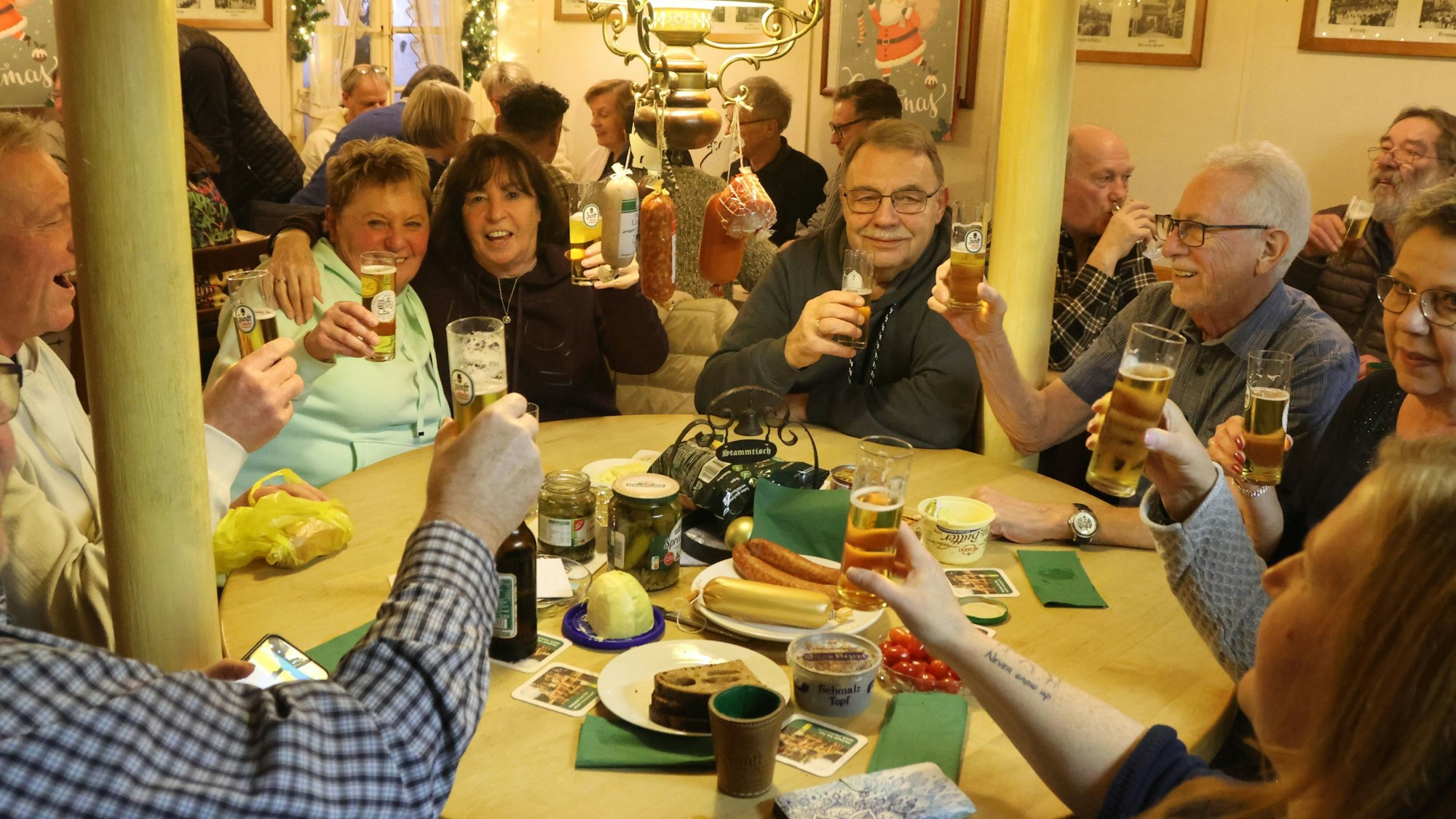 Zu sehen sind Menschen, die in einer Kneipe an einem Tisch ein Würfelspiel spielen. An der Lampe hängen Würste.