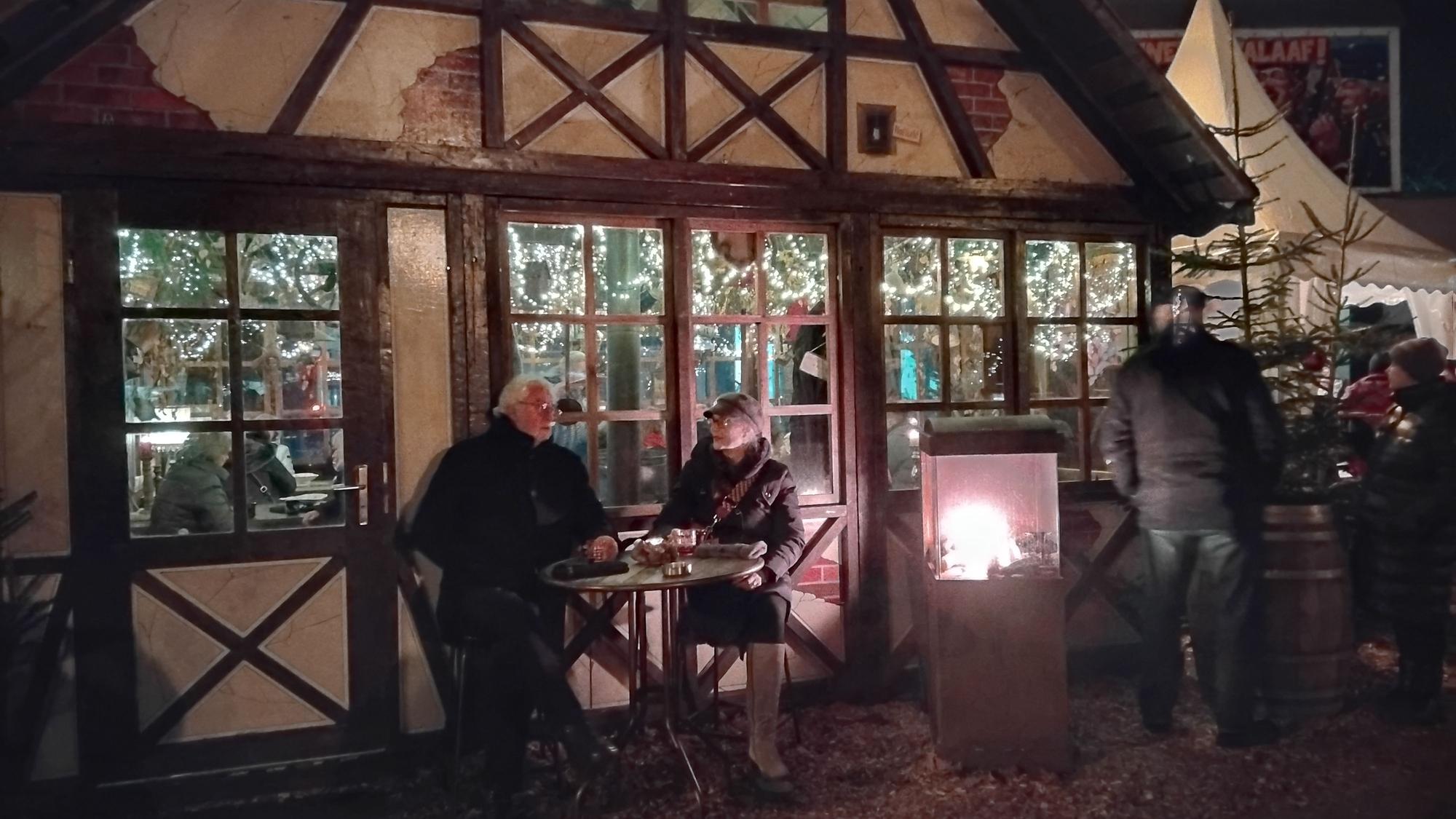 Eine beleuchtete Fachwerkhütte und ein Pagodenzelt, davor eine Feuerstelle und Menschen
