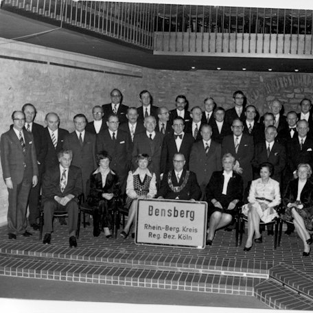 Die Mitglieder des Stadtrates der Stadt Bensberg stehen am 18. Dezember 1974 während ihrer letzten Sitzung mit einem Bensberger Ortsschild im Ratssaal des Bensberger Rathauses.