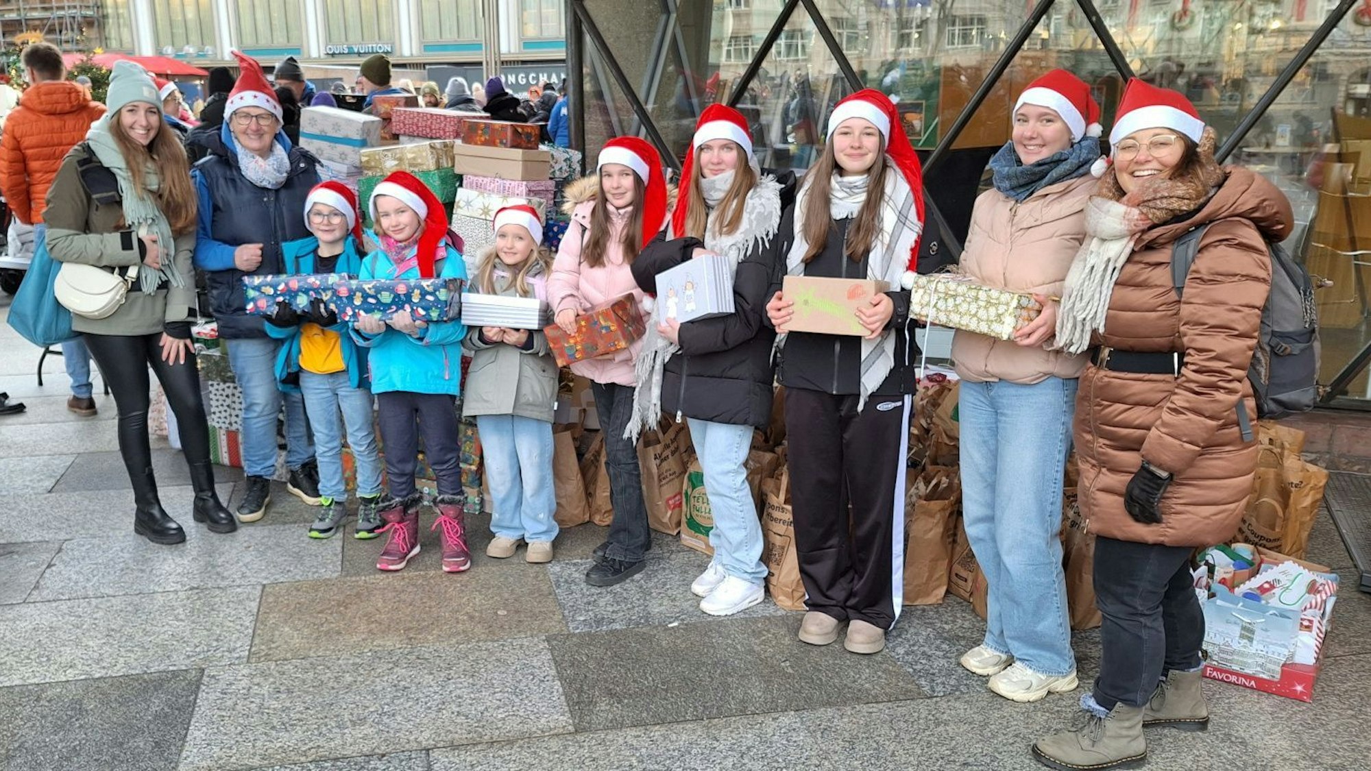 Kleine Kinder, Jugendliche und Erwachsene stehen auf der Domplatte in Köln. Sie tragen Weihnachtsmützen und halten verpackte Geschenke in die Kamera.