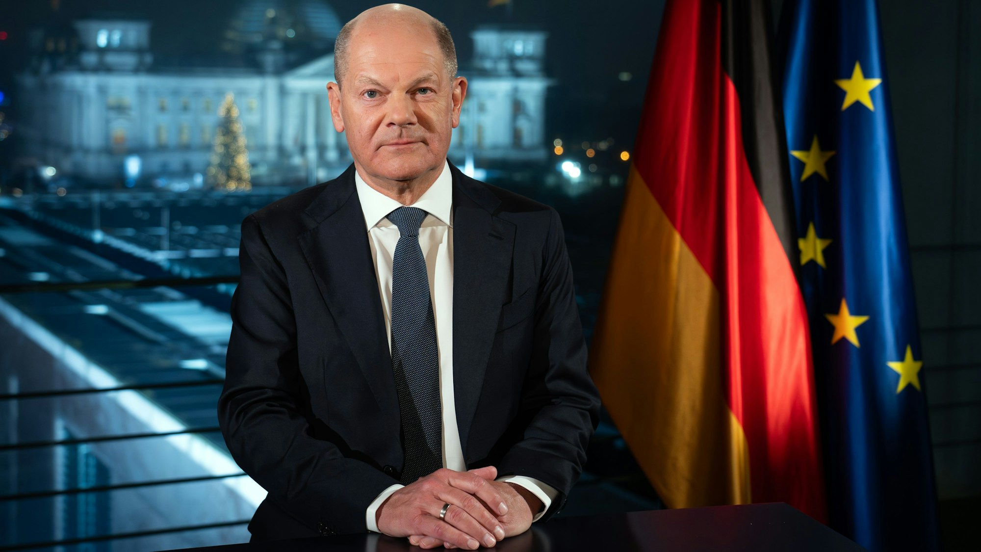 Bundeskanzler Olaf Scholz (SPD) sitzt während eines Fototermins nach der Aufzeichnung seiner Neujahrsansprache im Kanzleramt vor der Kulisse des von Scheinwerfern angestrahlten Reichstages.