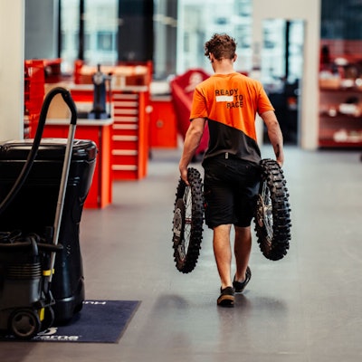 Bild eines Mannes, der ein Paar Motorradreifen durch eine Halle trägt.