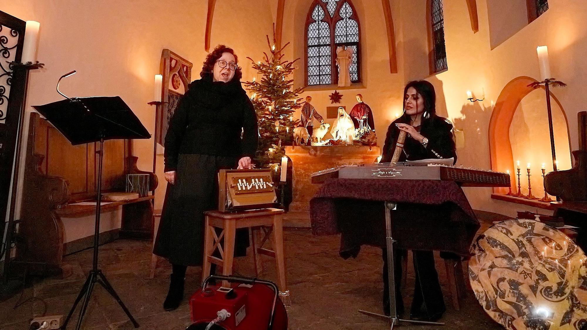 Die Musikerinnen Beate Alsdorf und Arezoo Resvani stehen in der von Kerzen erleuchteten Ahekapelle bei Engelgau im Altarraum, im Hintergrund ist ein Weihnachtsbaum.