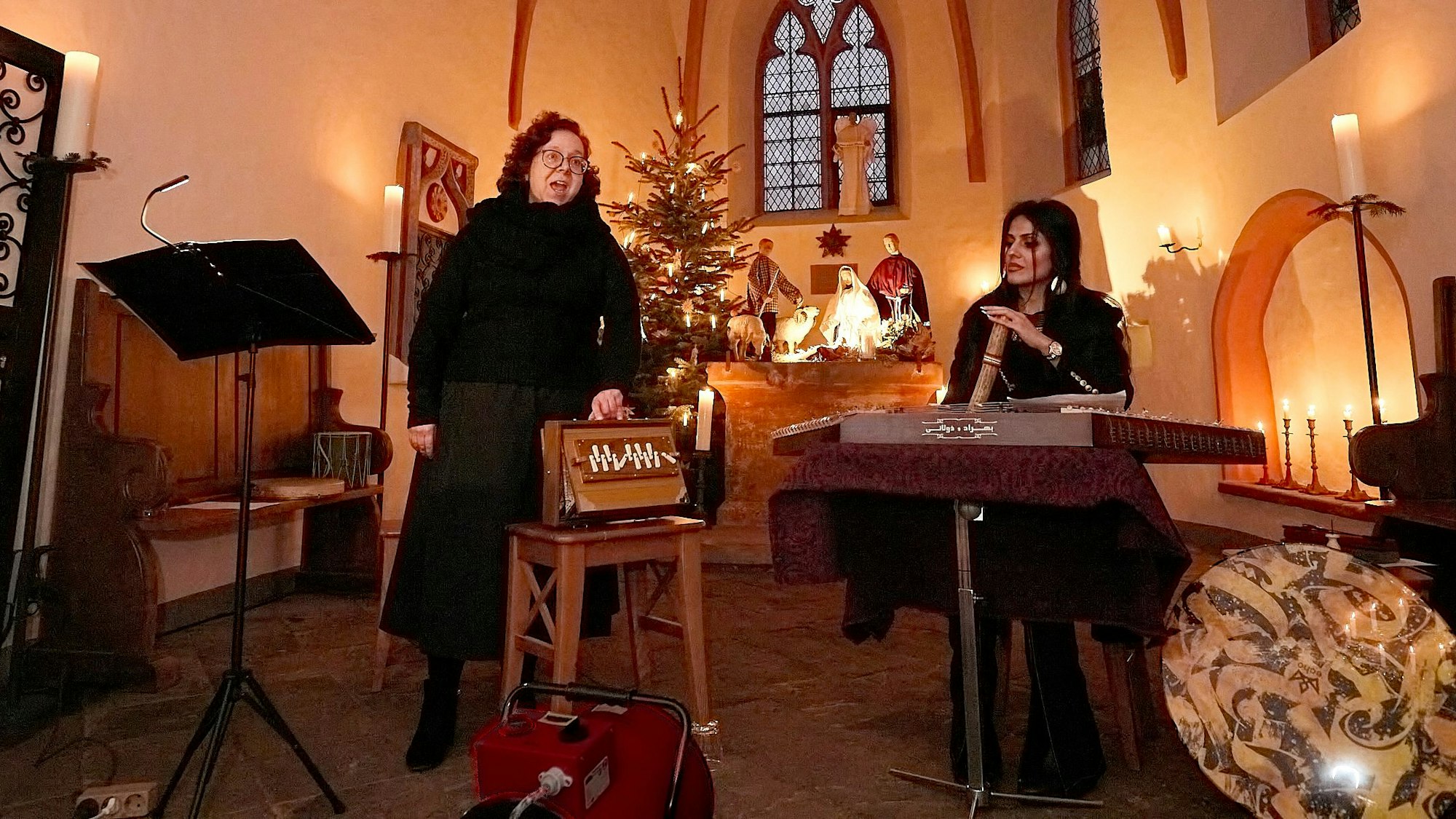 Die Musikerinnen Beate Alsdorf und Arezoo Resvani stehen in der von Kerzen erleuchteten Ahekapelle bei Engelgau im Altarraum, im Hintergrund ist ein Weihnachtsbaum.