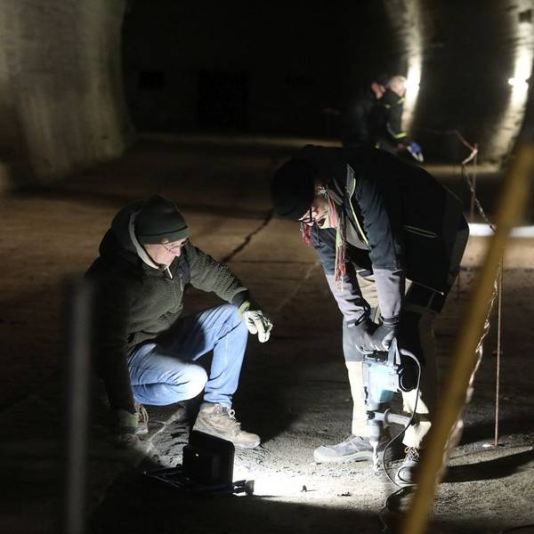 Zwei Männer mit einer Bohrmaschine in einem nur punktuell beleuteten Tunnel.