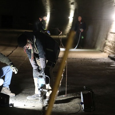 Zwei Männer mit einer Bohrmaschine in einem nur punktuell beleuteten Tunnel.