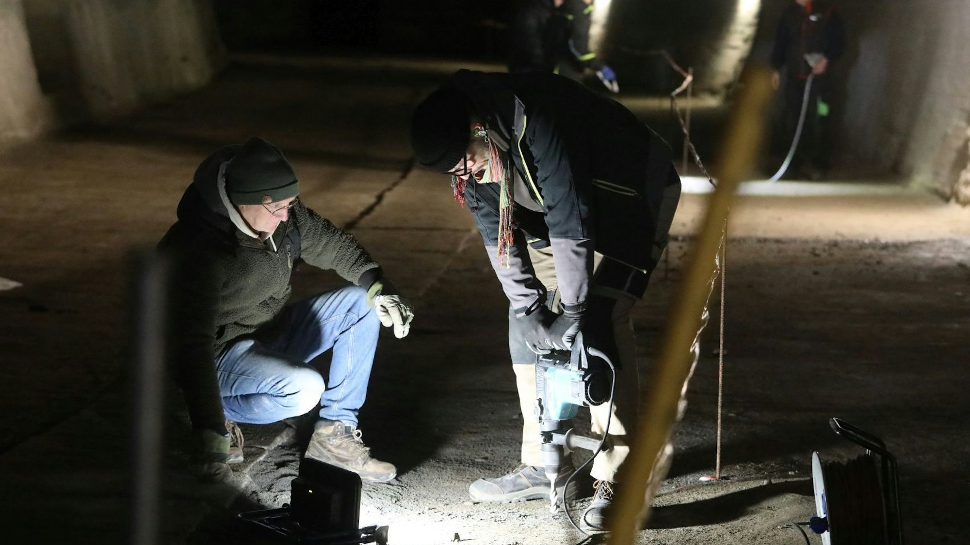 Zwei Männer mit einer Bohrmaschine in einem nur punktuell beleuteten Tunnel.