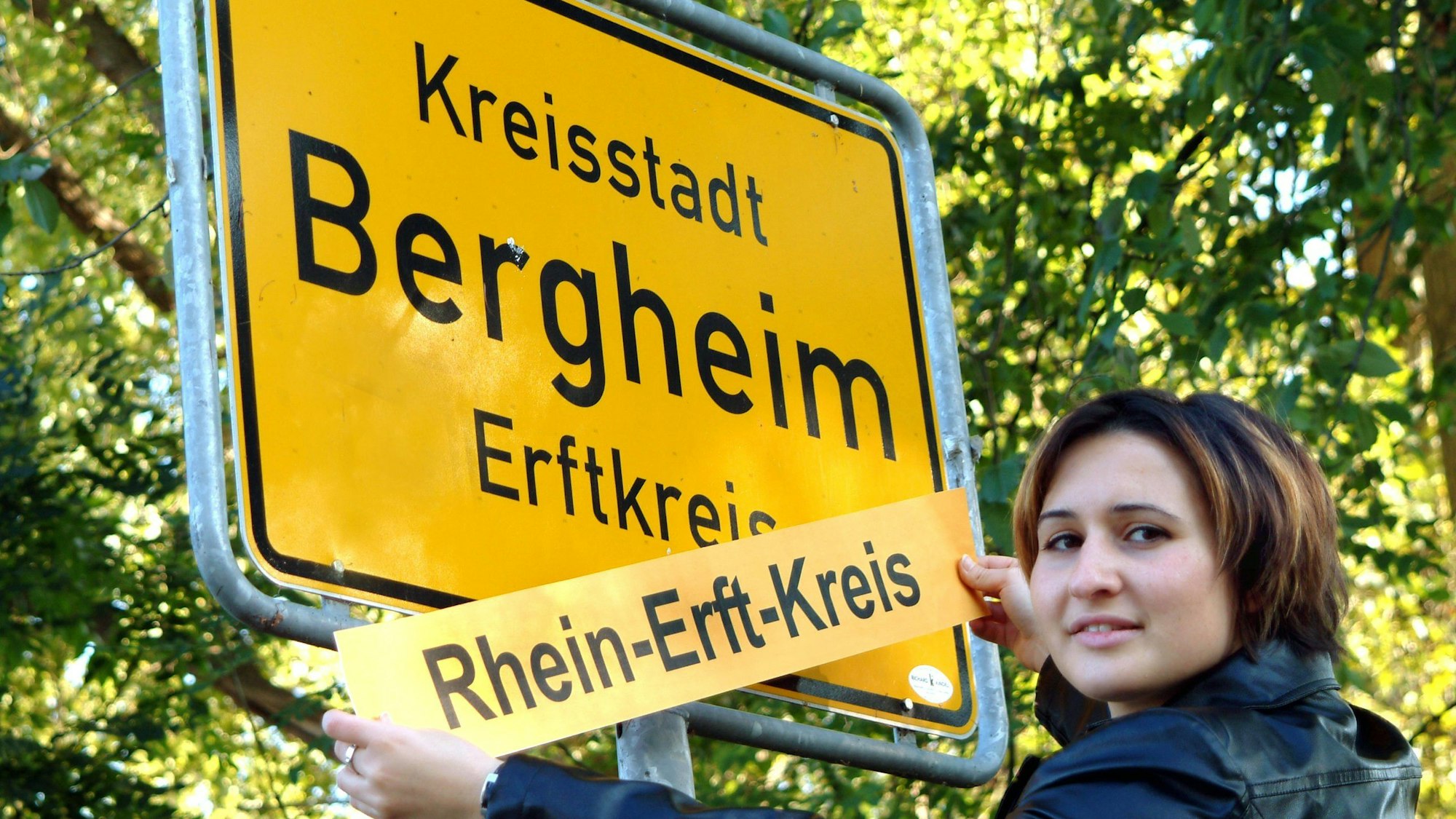 Das Bild zeigt eine Frau, die das Ortseingangsschild mit dem Aufkleber Rhein-Erft-Kreis überklebt.