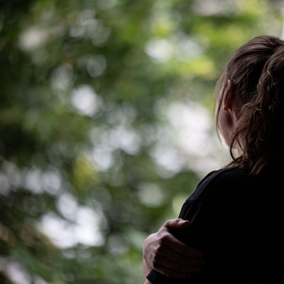 Eine Frau mit Pferdeschwanz steht mit dem Rücken zum Betrachther an einem geöffneten Fenster und schaut hinaus.