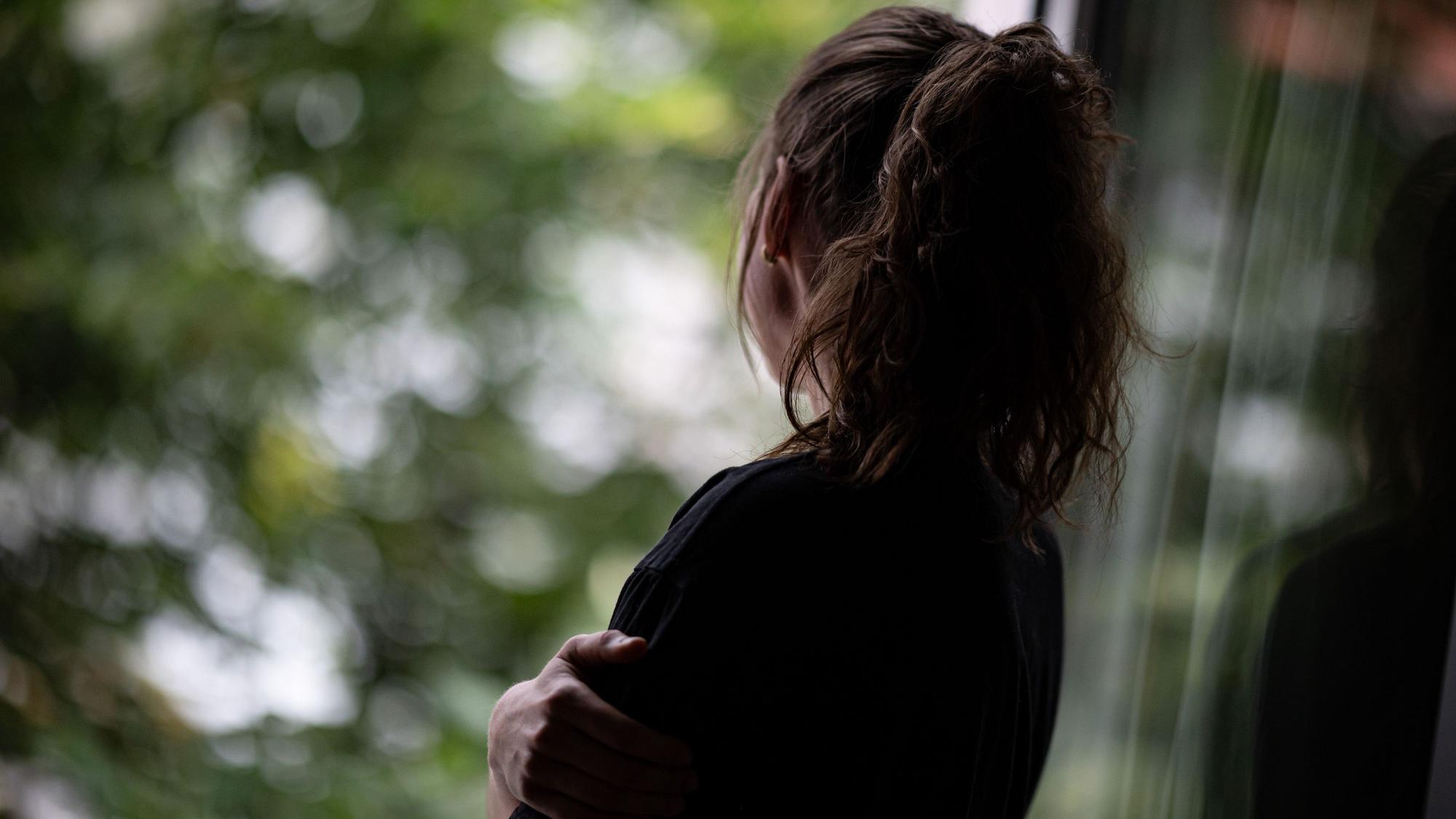 Eine Frau mit Pferdeschwanz steht mit dem Rücken zum Betrachther an einem geöffneten Fenster und schaut hinaus.