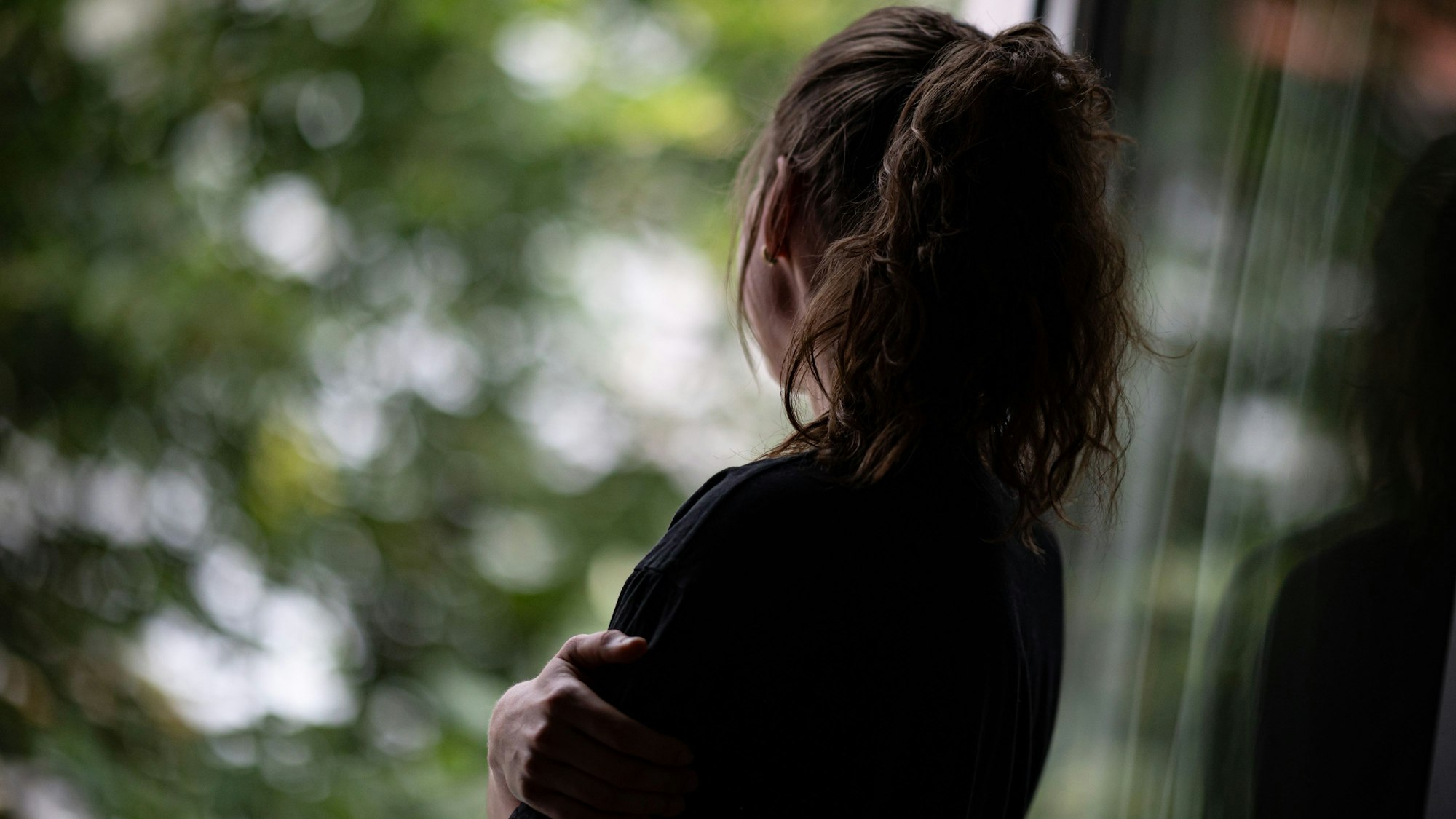 Eine Frau mit Pferdeschwanz steht mit dem Rücken zum Betrachther an einem geöffneten Fenster und schaut hinaus.