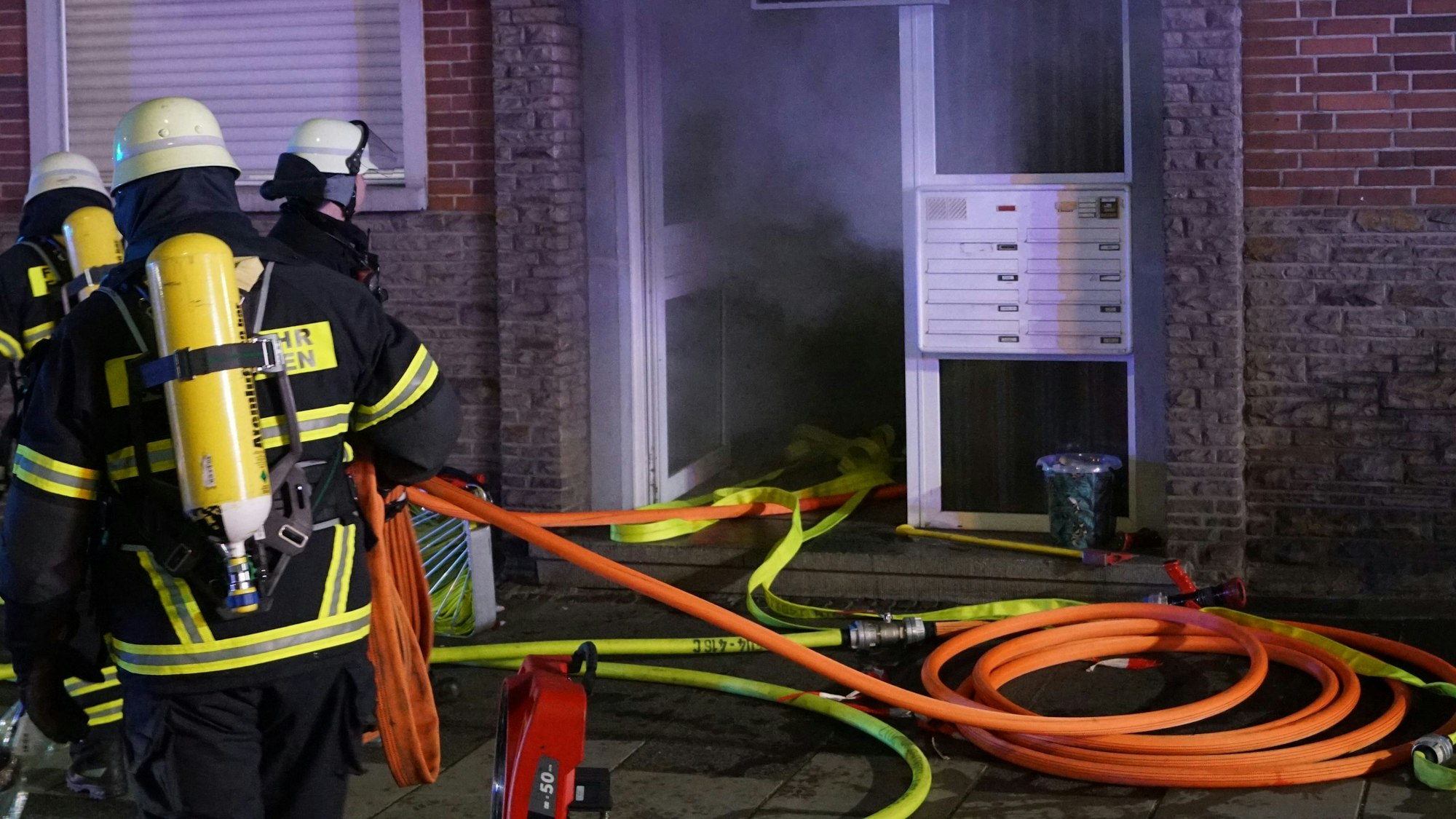 Drei Feuerwehrleute stehen vor einem Mehrfamilienhaus. Aus dem Eingang dringt Rauch. Auf dem Boden liegen gelbe und orangefarbene Schläuche.