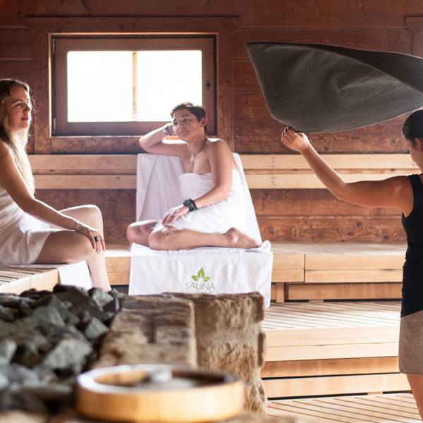 Zwei Frauen sitzen in der Sauna, eine Mitarbeiterin wedelt mit einem Handtuch nach dem Aufguss.