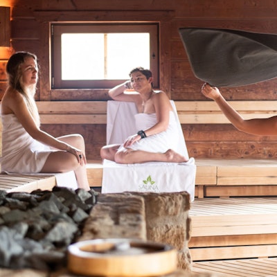 Zwei Frauen sitzen in der Sauna, eine Mitarbeiterin wedelt mit einem Handtuch nach dem Aufguss.