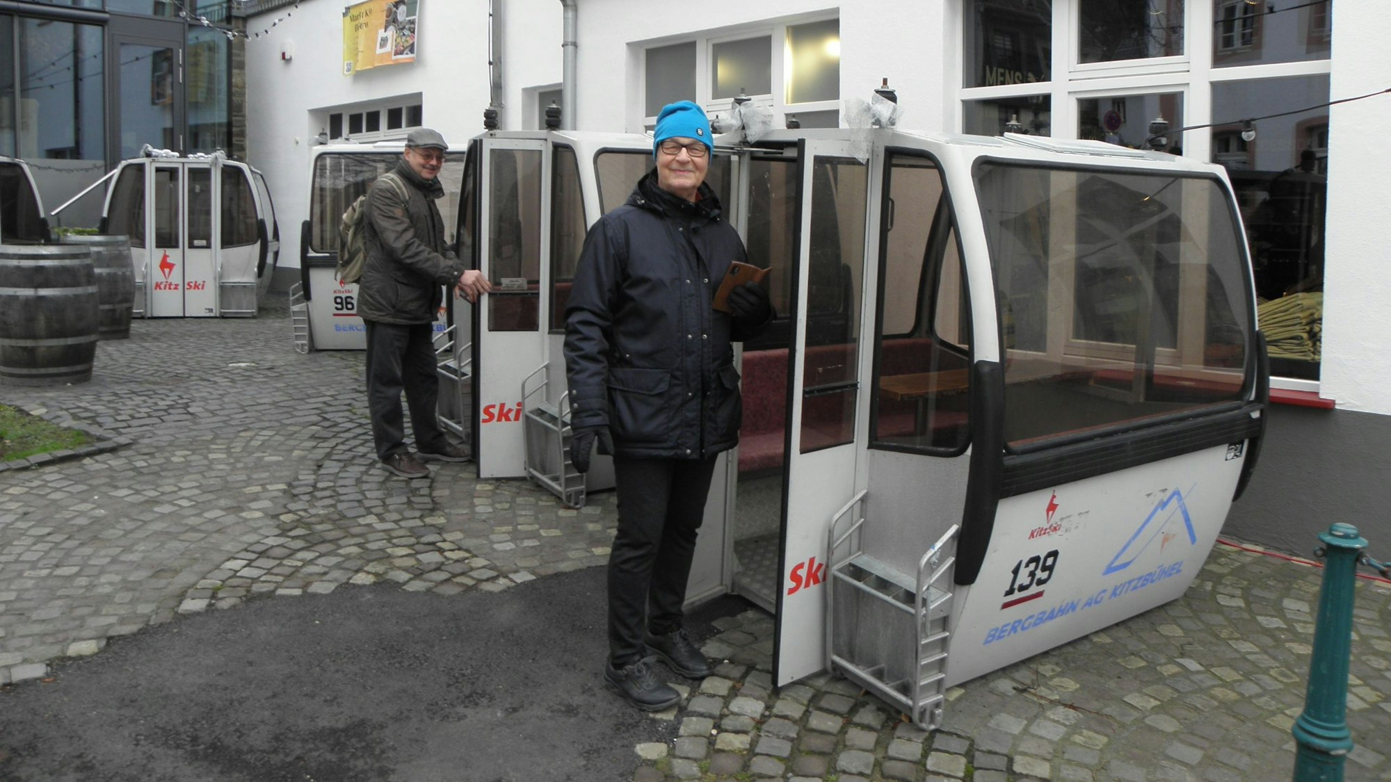 Mehrere Seilbahnkabinen stehen auf einem Parkplatz, ein Mann und eine Frau schauen sie interessiert an.