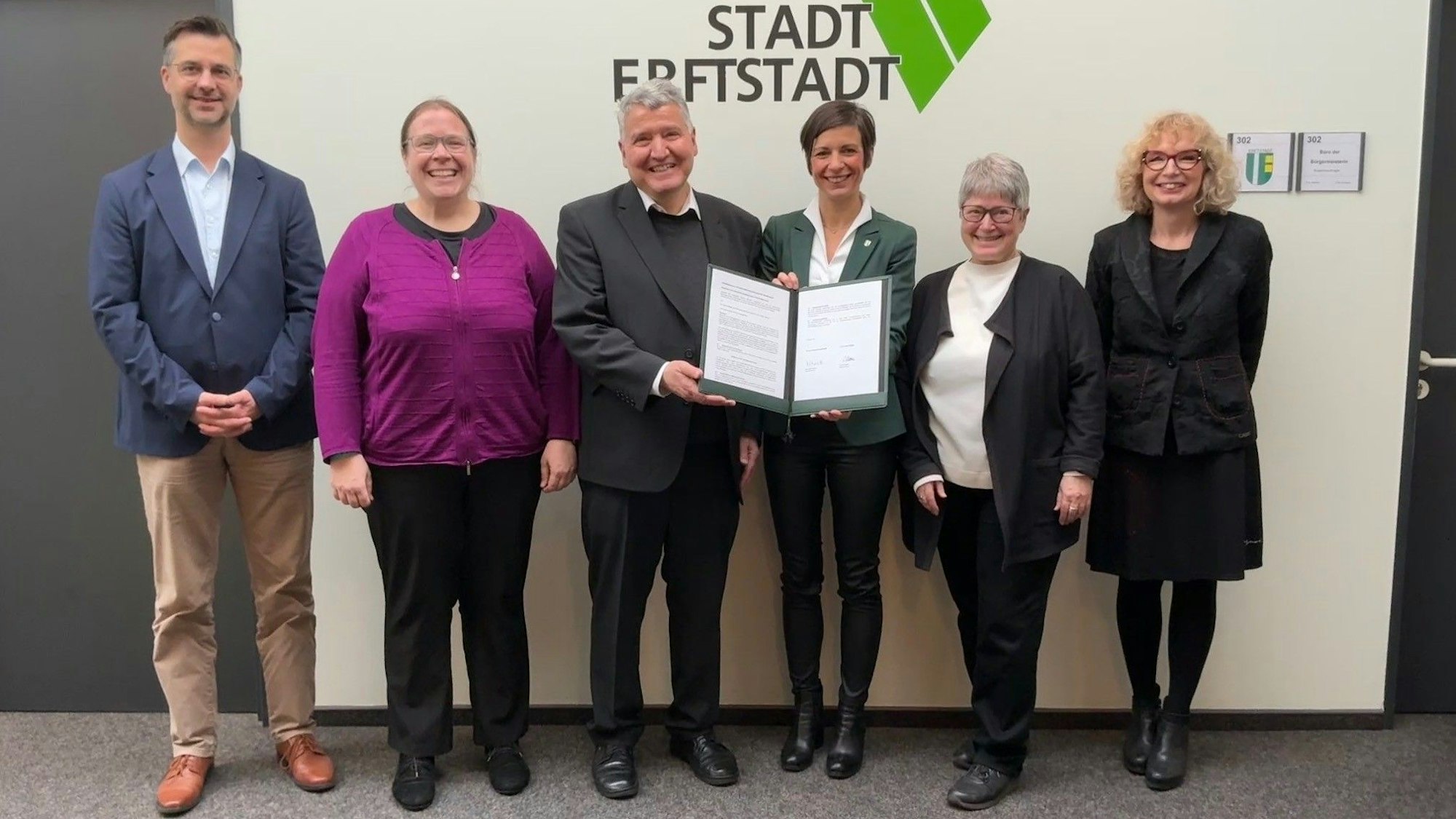 Männer und Frauen stehen vor einer Wand, zwei halten ein Dokument.