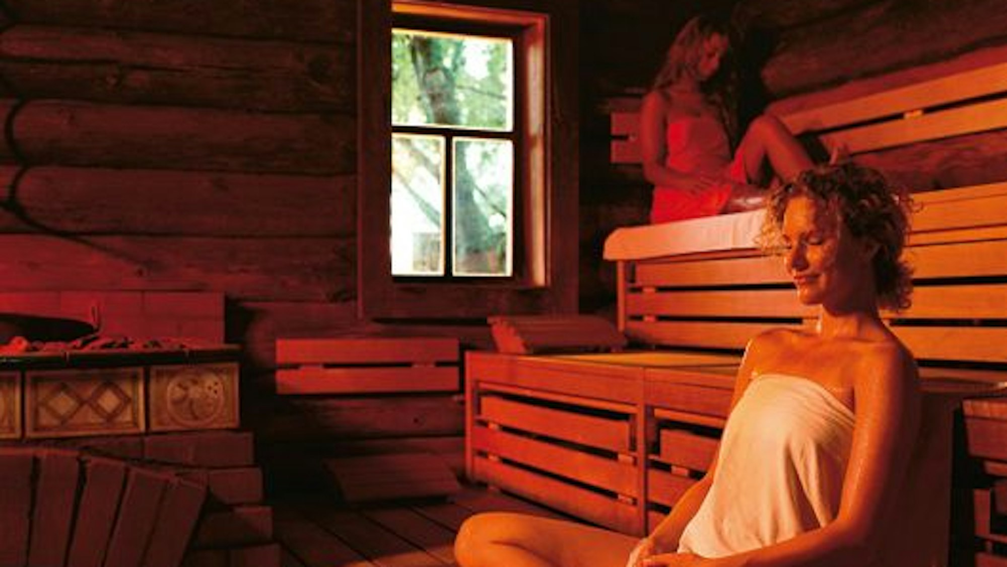 Zwei Frauen, die sich Handtücher umgewickelt haben, entspannen in einer Sauna, die rötlich beleuchtet wird.