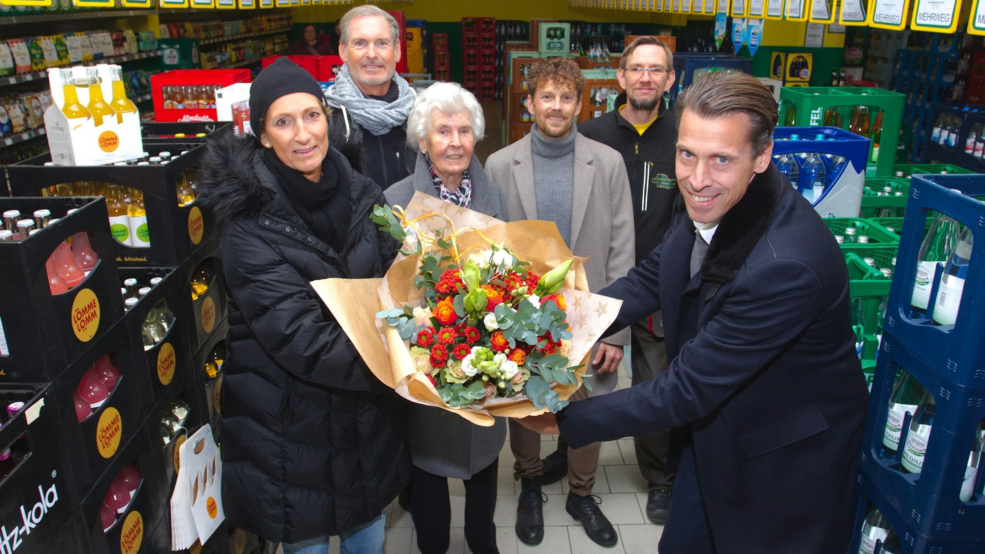 Ein Mann überreicht einer Frau im Beisein mehrerer weiterer Leute in einem Getränkemarkt einen großen Strauß Blumen.