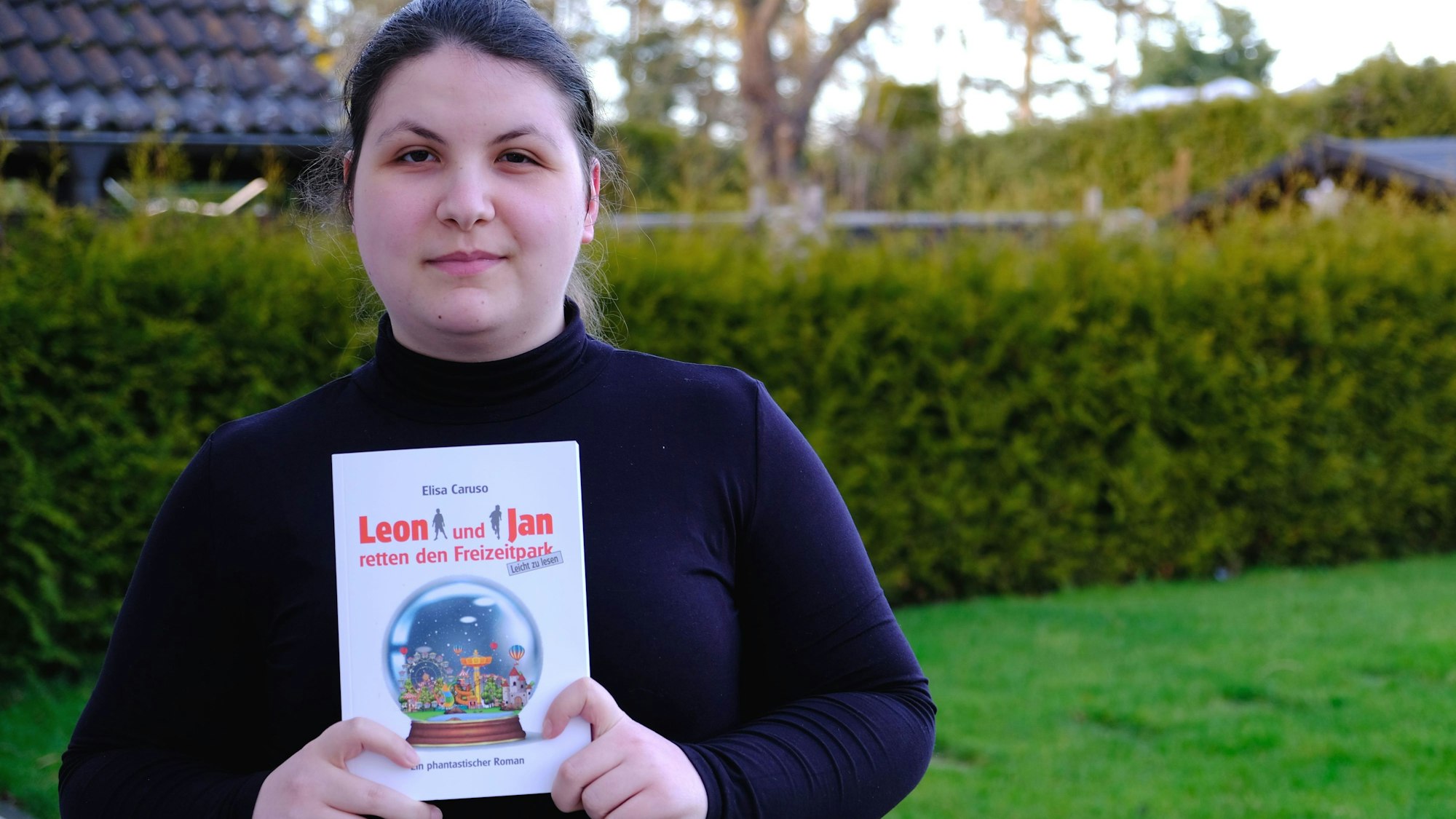 Elisa Caruso trägt einen schwarzen Rollkragenpulli und präsentiert ihr Buch mit dem Titel „Leon und Jan retten den Freizeitpark“.