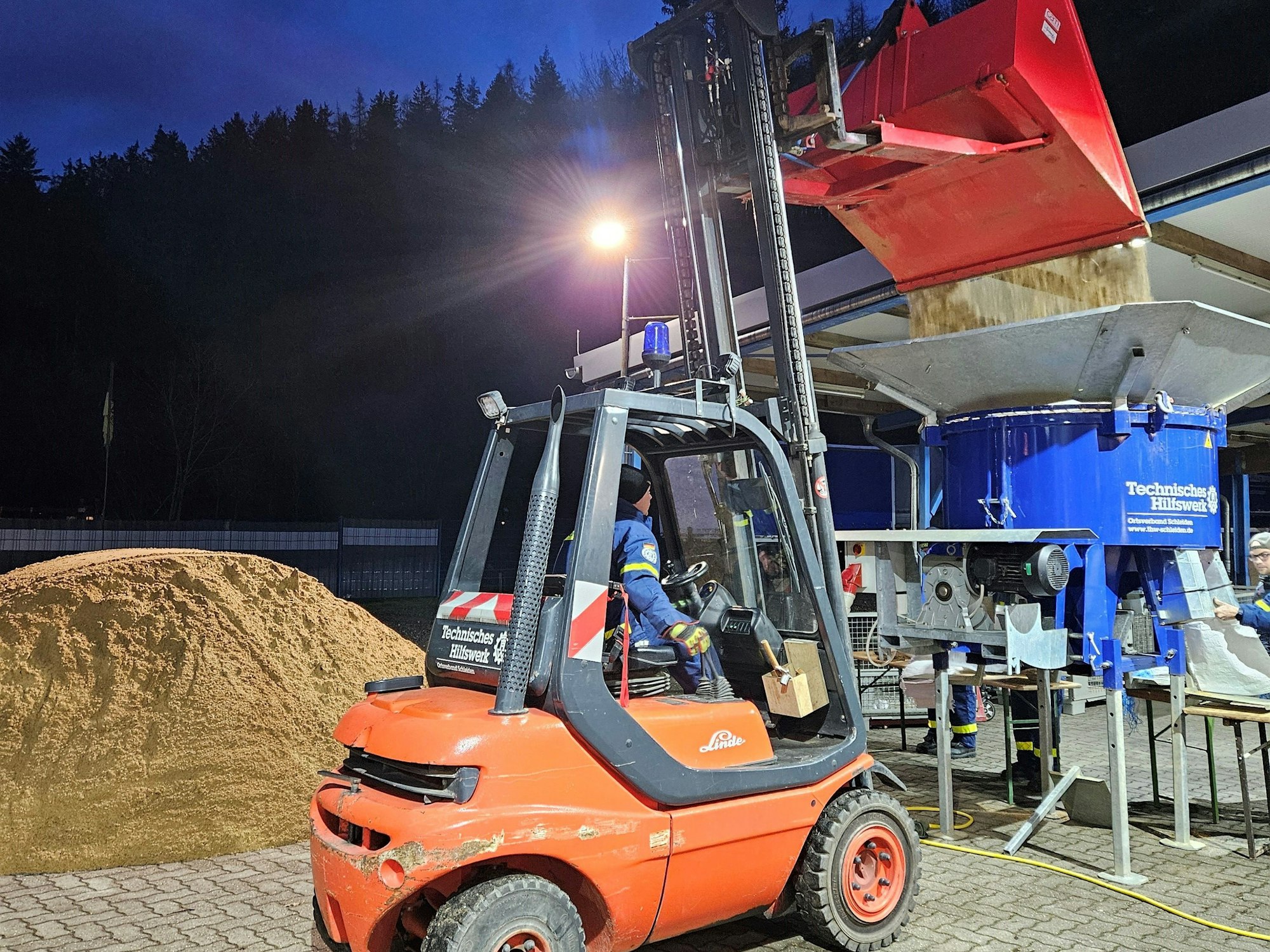 Das Bild zeigt einen Gabelstapler, der Sand in die Sandsackfüllmaschine kippt.