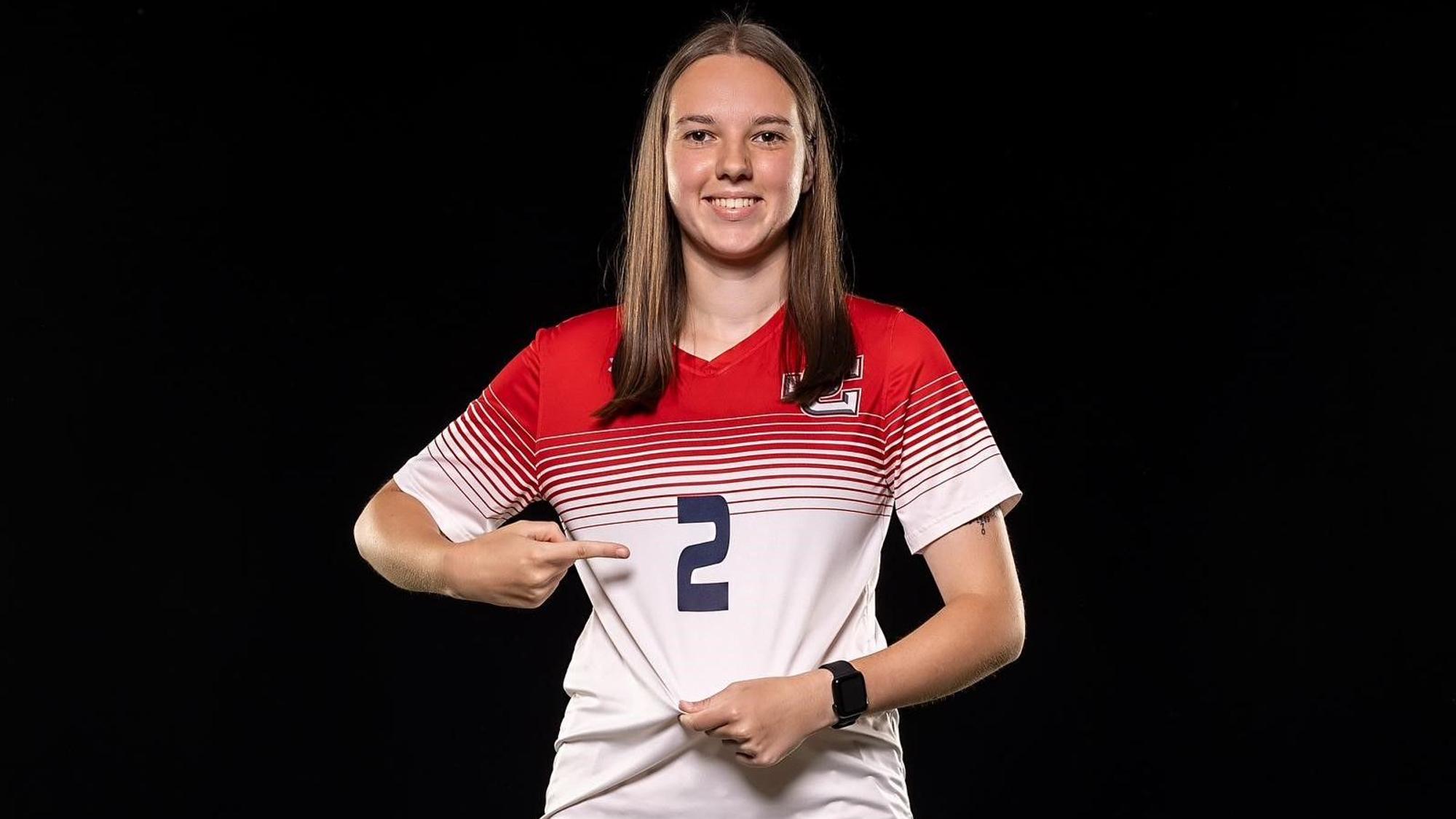 Lena Kücken trägt das Trikot mit der Nummer 2 des Jefferson College. Mit einem Finger zeigt sie auf die Nummer, mit der anderen Hand zieht sie das Trikot straff.