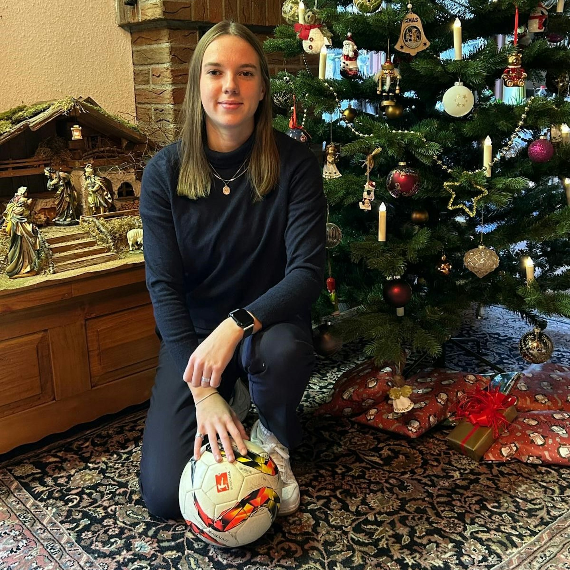 Lena Kücken hockt vor dem Weihnachtsbaum, unter dem Geschenke liegen, und vor einer Krippe. Ein Hand lehnt auf einen Fußball auf dem Teppichboden.