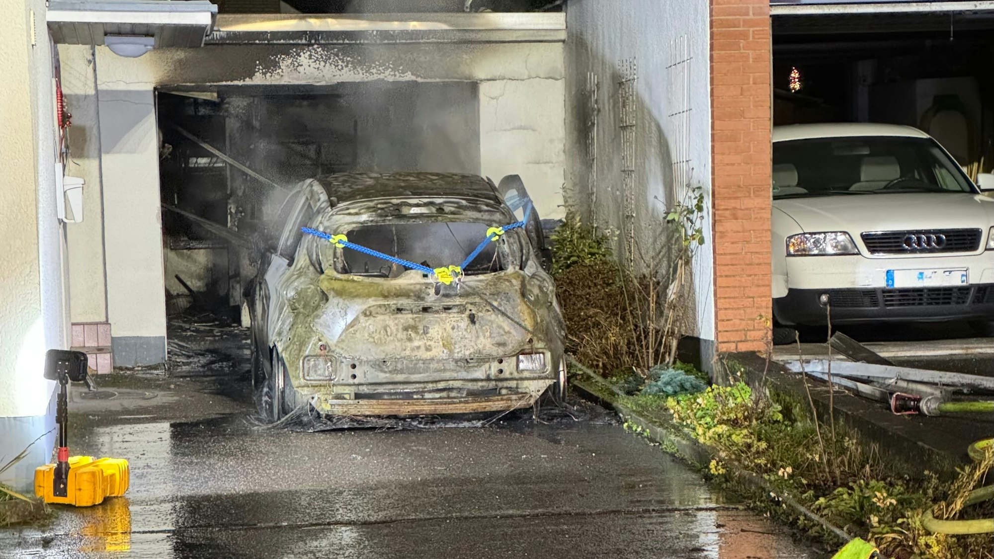 Ein völlig ausgebranntes Elektroauto wird von der Feuerwehr mit einer Seilwinde aus einer Garage am Neuenweg in Bergisch Gladbach gezogen.