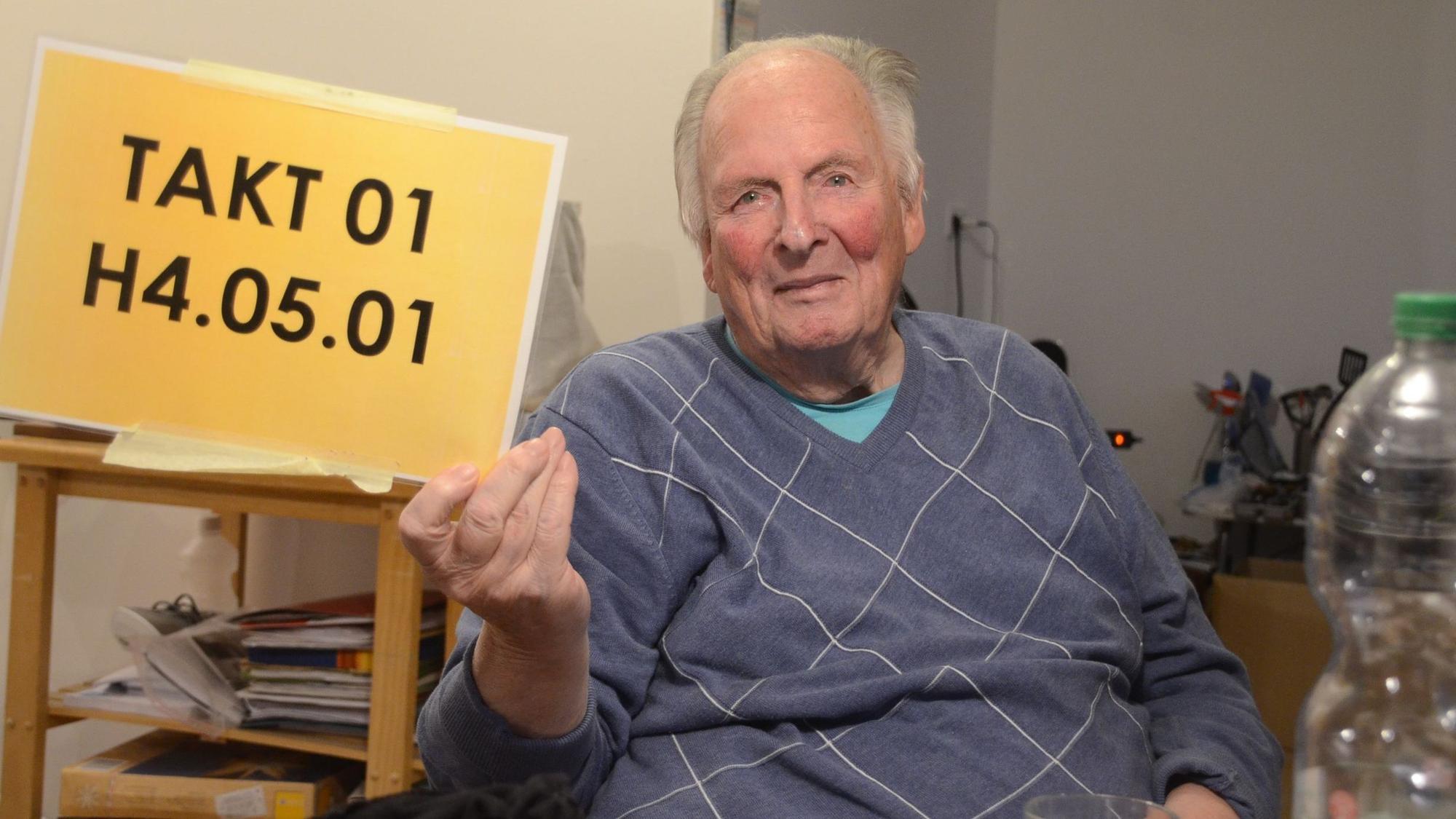 Ein Mann sitzt in einem Rollstuhl, er hält ein Schild mit einer Folge von Ziffern und Buchstabend hoch.