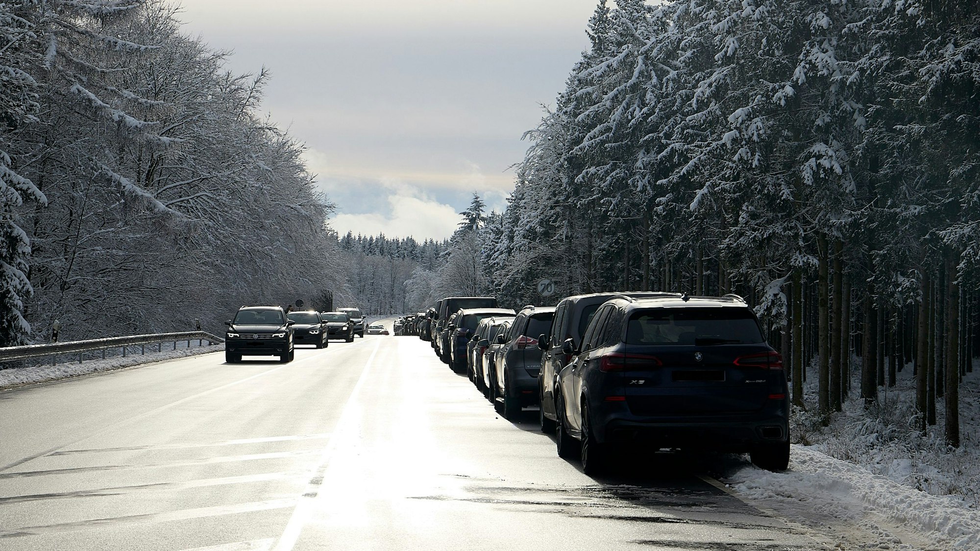 Eine lange Reihe von Autos ist am rechten Straßenrand der Bundestraße inmitten der verschneiten Fichten links und rechts geparkt.