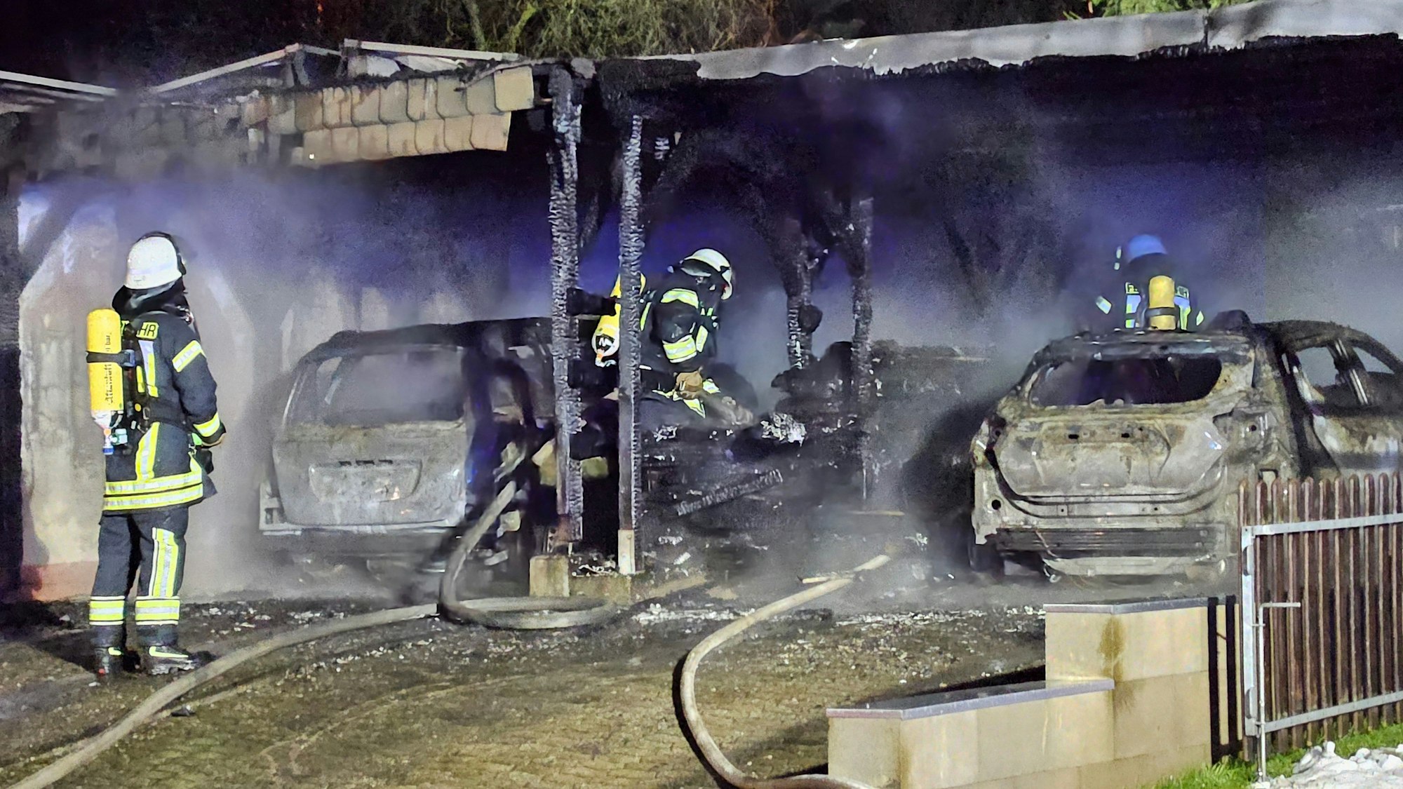 Feuerwehrleute begutachten nach dem Brand eines Carports zwei ausgebrannte Autowracks.