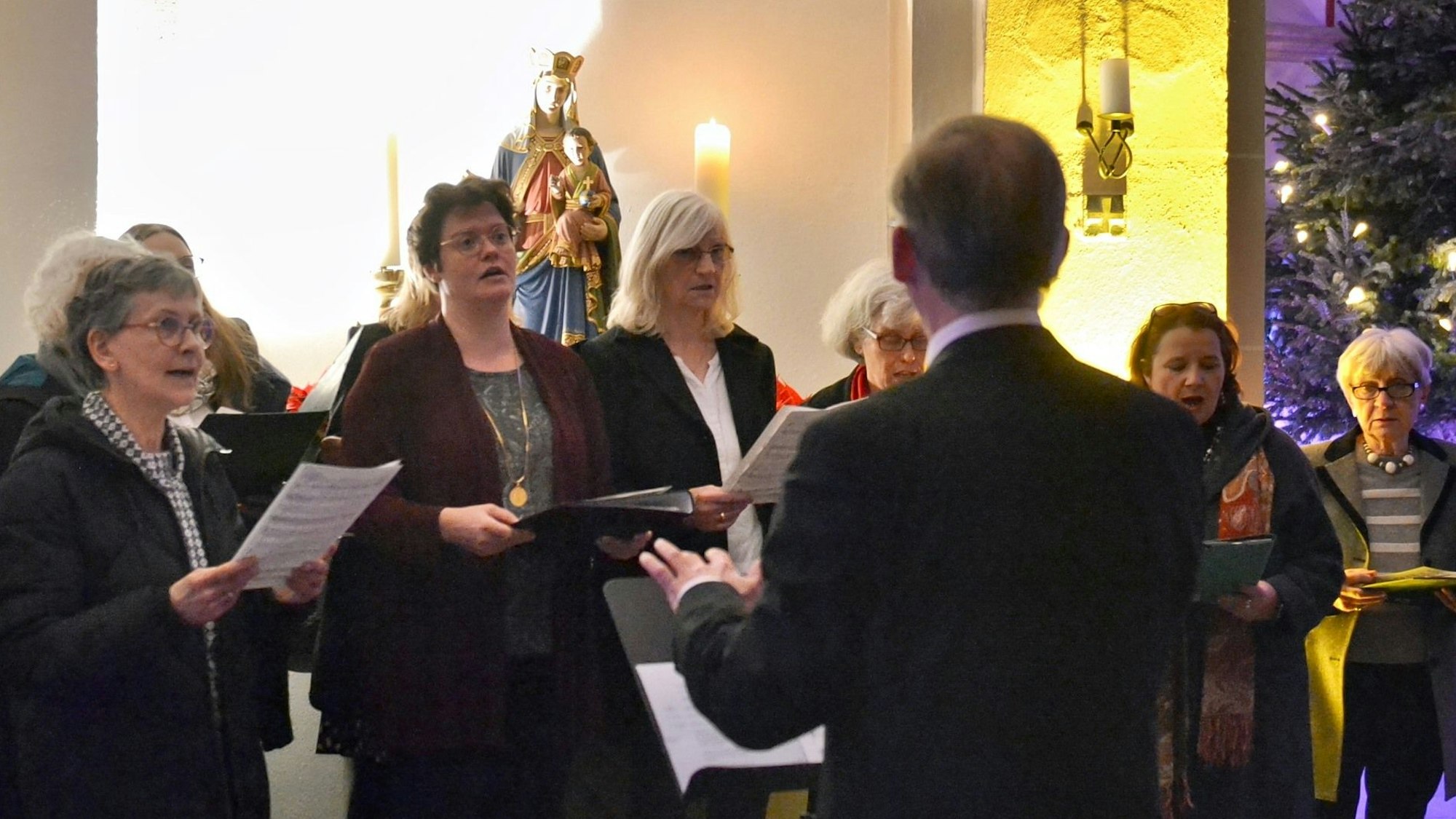 Mehrere Frauen halten Notenblätter in den Händen und singen, davor steht ein Dirigent, der nur von hinten zu sehen ist.