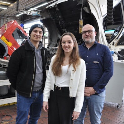 Auf dem Bild sind zwei Männer und eine junge Frau vor Lkws im Daimler Truck Nutzfahrzeugzentrums Mercedes-Benz in Frechen zu sehen.