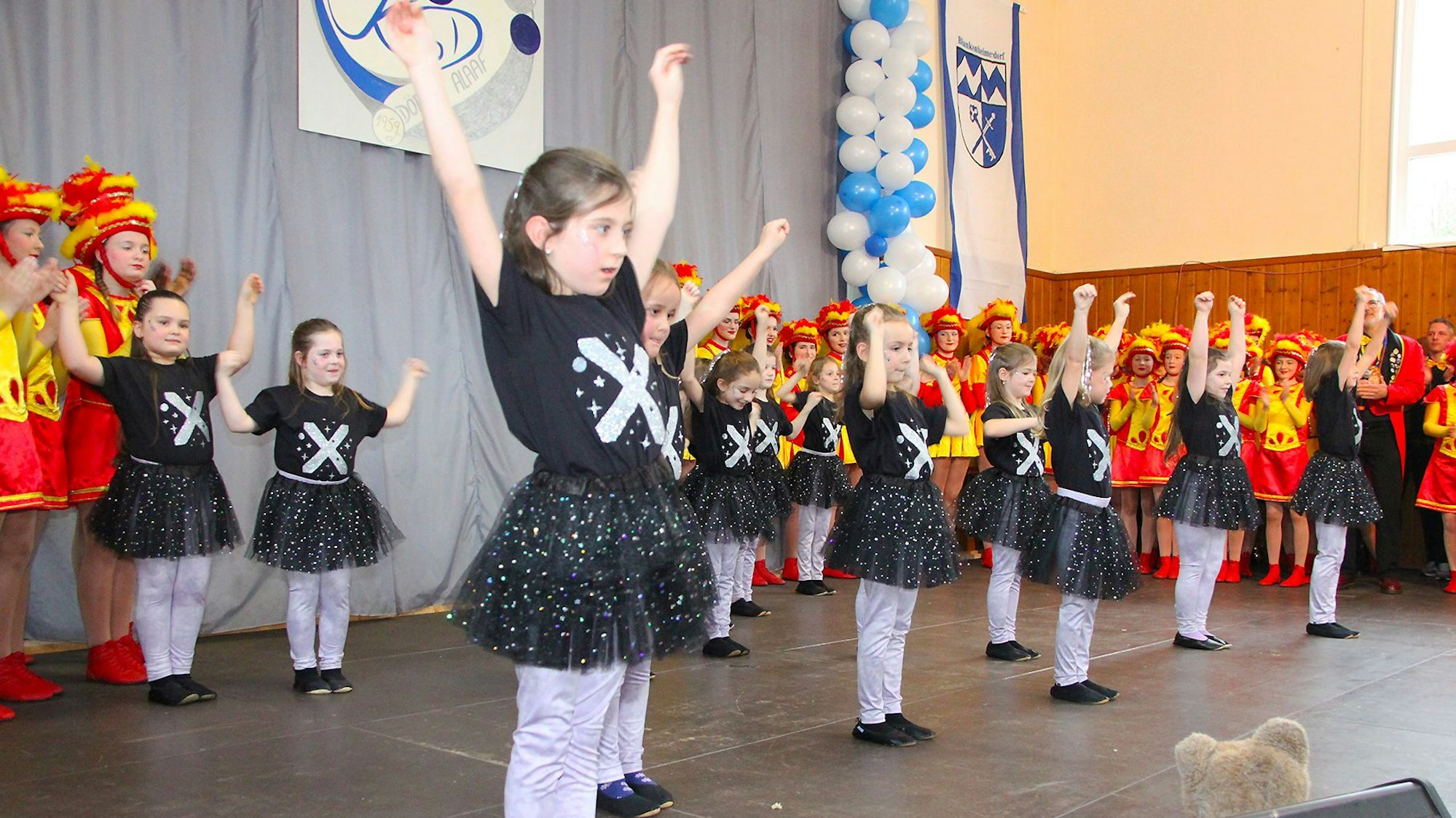 Eine Gruppe Kinder in schwarz-grauen Kostümen steht auf der Bühne und zeigt einen Tanz. Im Hintergrund stehen klatschende Mitglieder eines Karnevalsvereins in rot-gelben Tanzmariechen-Kostümen.