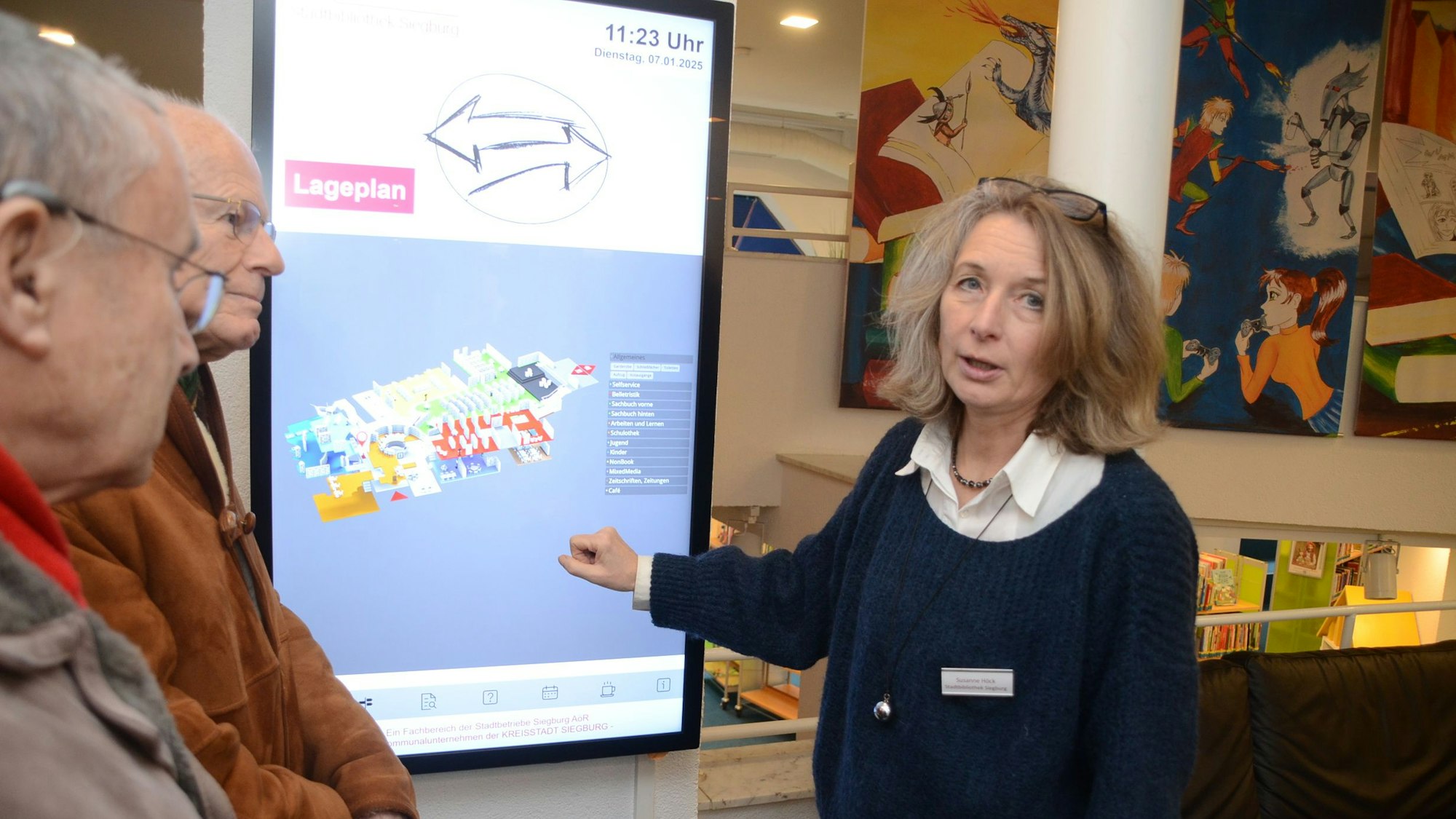 Eine Frau steht mit zwei Männern vor einem großen Bildschirm mit dem Lageplan einer Bibliothek.