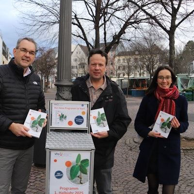 Zwei Männer und eine Frau stehen vor einem Kasten, aus dem Passanten das neue VHS-Programm entnehmen können.