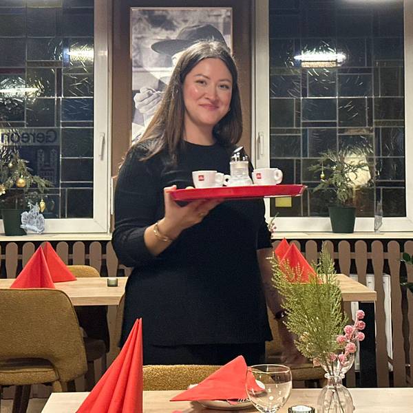 Eine Frau mit dunklen Haaren hält ein Tablett in der Hand, auf dem Espresso-Tassen stehen. Sie steht im Gastraum des Restaurants Buon Cibo in Köln-Zündorf.