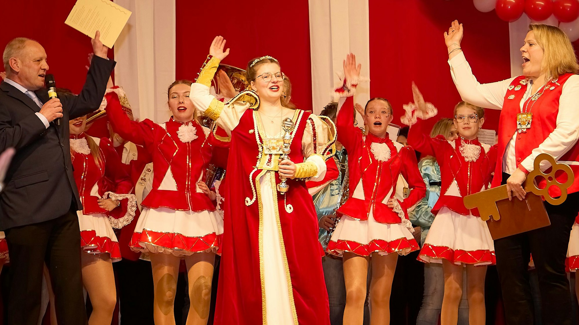 Karnevals-Prinzessin Ophelia I. steht im rot-weißen Ornat zusammen mit den Tänzerinnen einer Garde und weiteren Personen auf der Bühne; alle heben die Hand Karnevalsgruß.