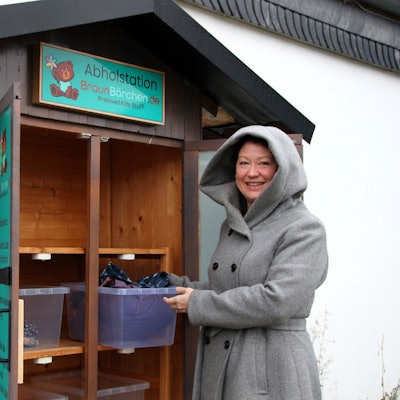 Sina Braun steht vor einem Außenschrank, in dem große Plastikboxen stehen. Sie trägt einen Mantel mit Kapuze und schaut in die Kamera.
